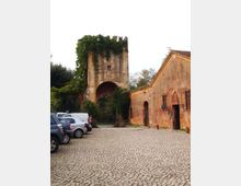 Cortile lastricato in ciottoli con auto parcheggiate, dominato da una torre in muratura antica con grande arco d’ingresso e vegetazione sulla sommità. Sulla destra si vede un lungo edificio rustico intonacato con tetto in coppi e porte ad arco.