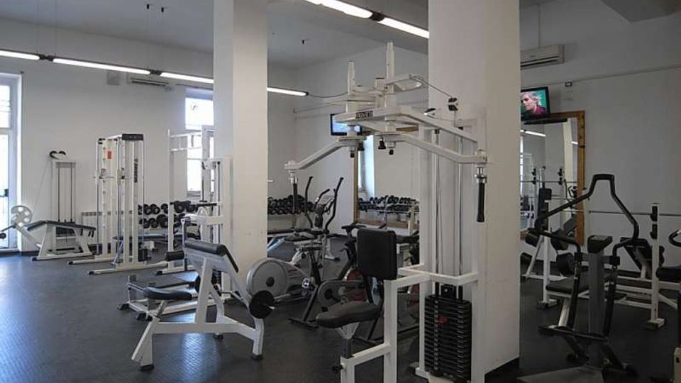 Interno di una palestra con ampie colonne bianche, illuminazione a neon e pavimento in gomma scura. Sono presenti diverse macchine per pesi e cardio, panche e una fila di manubri lungo la parete con grandi finestre.