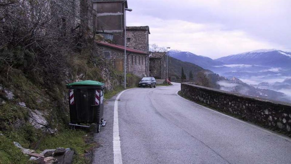 Strada asfaltata di montagna che curva lungo un muretto in pietra, con case in pietra addossate al pendio sul lato sinistro. Sullo sfondo si vedono vallate nebbiose e cime montuose, con un cassonetto verde in primo piano vicino al margine della strada.