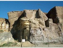 Resti di una fortificazione in pietra con muri spessi e irregolari, addossata a una parete rocciosa chiara. Davanti si vede un’area arida recintata con pali e un piccolo cartello informativo sotto un cielo sereno.