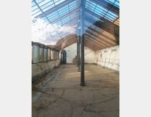 Interno di una grande serra o capannone abbandonato con tetto a falde in vetro e struttura metallica, sostenuto da colonne in ferro. Pavimento in cemento con crepe e vegetazione, pareti laterali con finestre e mensole metalliche, e un arco d’ingresso sul fondo.