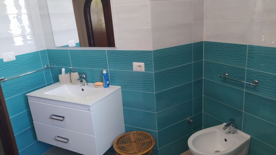 Bagno moderno con pareti piastrellate bianche e fascia turchese, lavabo con mobile sospeso e grande specchio. A destra si vede un bidet e una barra portasciugamani, con piccolo sgabello in vimini sul pavimento.