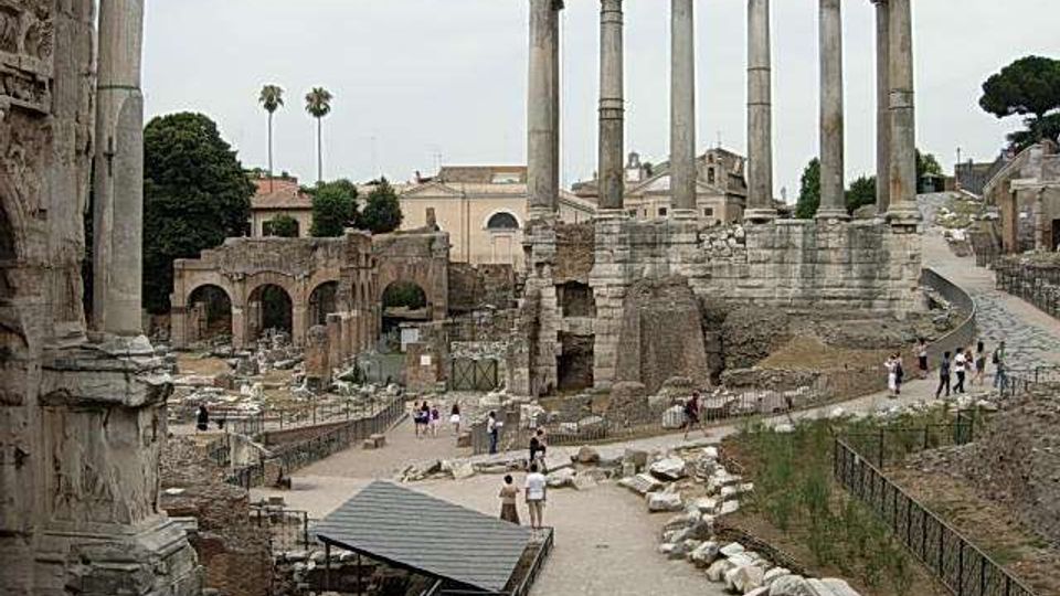 Rovine di un sito archeologico romano con un tempio su alto basamento e alte colonne in pietra, circondato da resti di mura e archi. Sentieri sterrati e recinzioni attraversano l’area con blocchi di pietra sparsi, con edifici e alberi sullo sfondo.