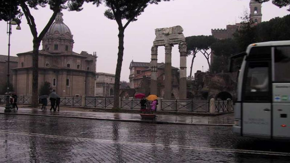 Veduta urbana in una giornata piovosa con strada in sampietrini bagnati in primo piano, alberi ad alto fusto e una recinzione lungo il marciapiede. Sullo sfondo si vedono rovine archeologiche con colonne e un arco, affiancate da una chiesa con grande cupola.