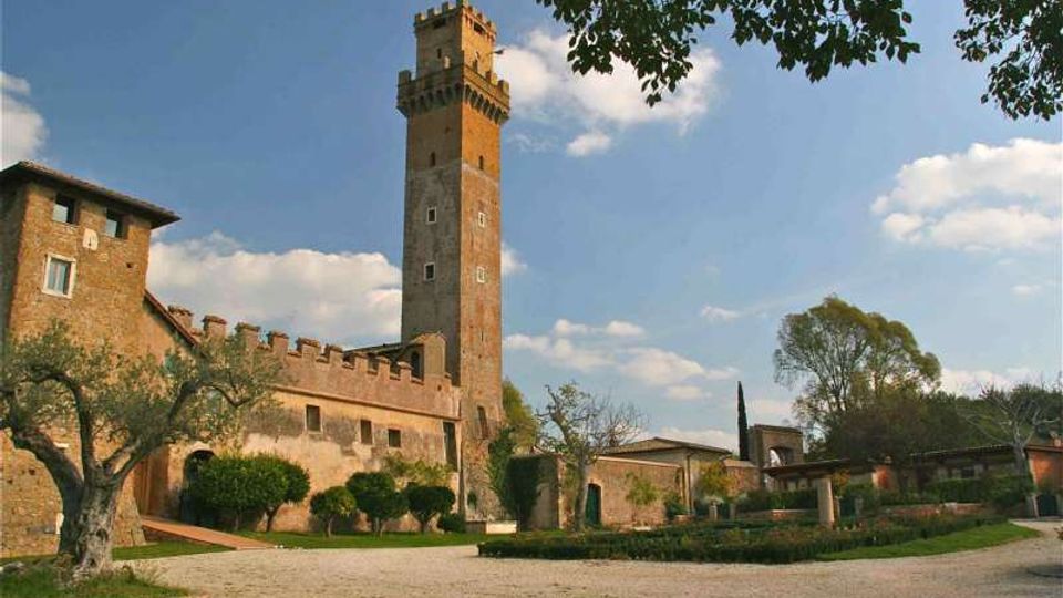 Castello in pietra e mattoni con alta torre merlata, affiancato da mura con camminamenti e archi. Davanti si estende un cortile ghiaioso con giardini curati e alberi, sotto un cielo azzurro con nuvole.