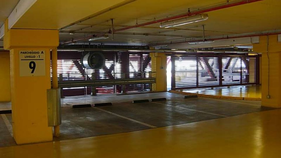 Interno di un parcheggio multipiano con pilastri e soffitto gialli, tubazioni e lampade a vista, e posti auto delimitati da strisce bianche. Sul pilastro è presente un cartello “Parcheggio A Livello -1” con il numero 9 e sul fondo si vedono pannelli a griglia che lasciano filtrare la luce dall’esterno.