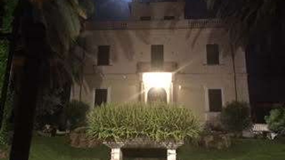 Edificio storico a due piani illuminato di notte, con ingresso centrale ad arco e finestre simmetriche sulla facciata. Davanti si vede un giardino con prato, aiuola rialzata e palme ai lati, sotto un lampione.