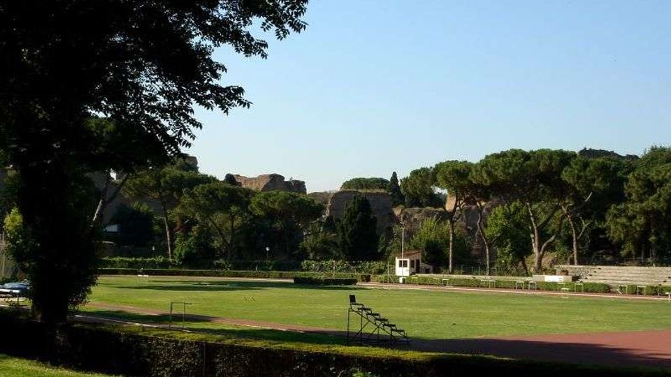 Campo sportivo all’aperto con ampia area erbosa centrale e pista ovale rossa attorno, con una piccola tribuna metallica a bordo campo. Sullo sfondo si vedono alberi alti e colline con edifici in muratura, sotto un cielo sereno.