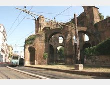 Ruderi di un antico acquedotto in mattoni con grandi arcate sovrapposte, accanto a una strada urbana. In primo piano si vedono i binari e la linea elettrica del tram, con edifici ai lati e vegetazione che cresce sulle mura.