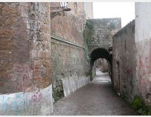 Vicolo stretto in pietra con pavimentazione in ciottoli, fiancheggiato da alti muri in muratura grezza con tubazioni a vista e tracce di graffiti. In fondo si vede un arco in pietra che forma un passaggio coperto verso una piccola curva della strada.