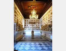 Interno di una galleria museale con soffitto in legno a cassettoni e grande lampadario centrale, pavimento in marmo a motivi geometrici. Lungo le pareti bianche con riquadri decorativi sono disposti numerosi busti e statue su mensole, con un piedistallo al centro della sala.