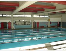 Piscina coperta con più corsie delimitate da galleggianti, vasca rettangolare e bordi piastrellati. L’ambiente presenta grandi finestre lungo le pareti, porte rosse e tubazioni rosse a vista sul soffitto con travi bianche.