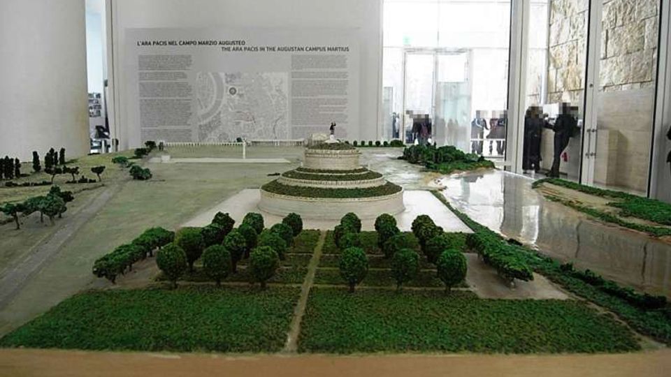 Modello in scala di un parco archeologico con alberi in miniatura, vialetti e una struttura circolare terrazzata al centro. L’allestimento è in una sala museale luminosa con pareti bianche, pannelli informativi e grandi vetrate sul lato destro.