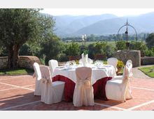 Tavolo rotondo apparecchiato all’aperto su una terrazza in mattoni, con sedie rivestite e tovaglia bianca e rossa. Sullo sfondo si vedono un giardino con alberi e vasi, un pozzo in pietra e una valle con colline e montagne.