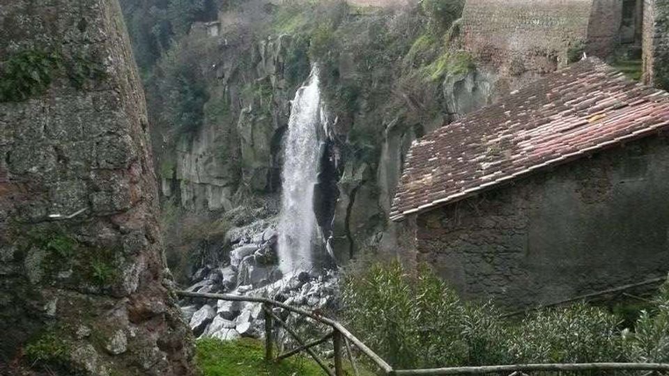 Veduta di una gola rocciosa con una cascata che scende tra pareti di pietra, vista da un sentiero erboso con staccionata in legno. Ai lati si notano murature in pietra e un edificio con tetto in tegole, con vegetazione sulle scarpate e costruzioni in lontananza.
