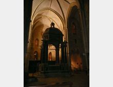 Interno di una chiesa in pietra con alte volte a crociera e archi gotici; al centro si trova un’edicola o altare coperto con piccola cupola e croce, circondato da colonne. Sullo sfondo si vedono finestre strette con vetrate scure e illuminazione soffusa.