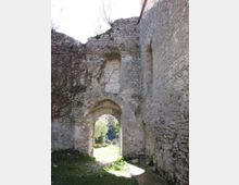 Rovine di una costruzione in pietra con alte mura e un arco d’ingresso, con resti di intonaco e mattoni visibili. Attraverso l’arco si vede un’area erbosa illuminata, circondata da vegetazione e alberi.