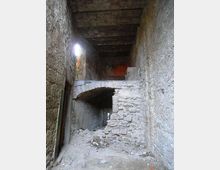 Interno di un edificio in pietra in rovina, con un arco in muratura parzialmente crollato e macerie sul pavimento. Pareti e soffitto grezzi e danneggiati, illuminati da una piccola apertura laterale.