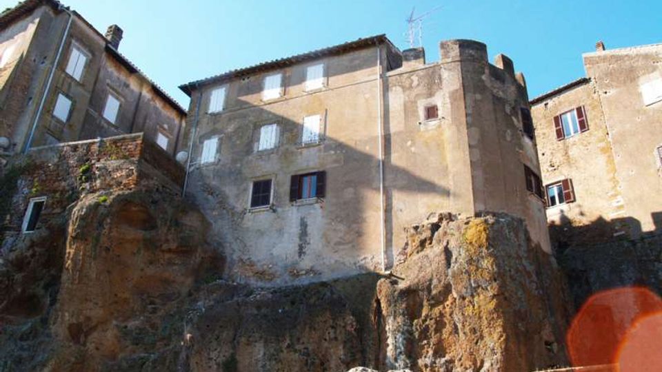Case in pietra e intonaco di un borgo storico costruite sopra alte rocce, con finestre e una torre cilindrica. In basso si vedono pareti rocciose e un piccolo tratto di strada o passaggio, sotto un cielo sereno.