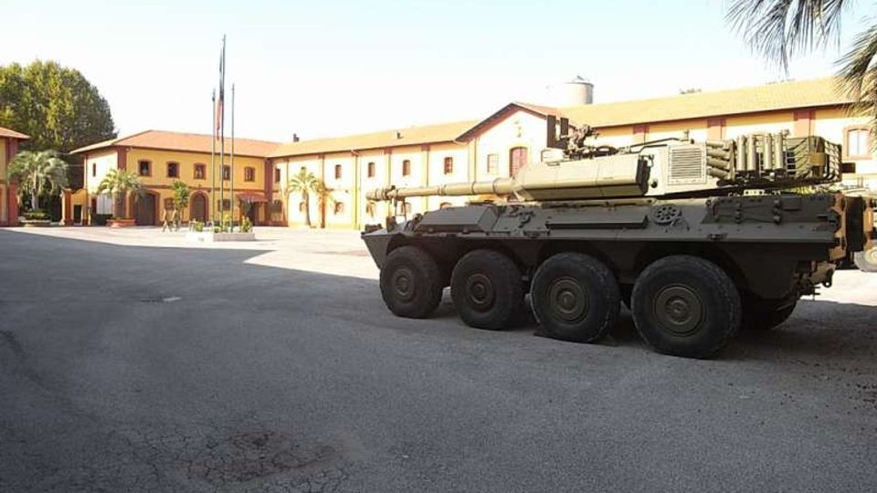 Piazzale asfaltato di una caserma con edifici bassi color ocra e tetti a falde sullo sfondo, con pennoni delle bandiere e palme. In primo piano è parcheggiato un veicolo militare blindato a otto ruote.
