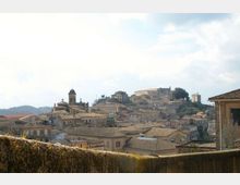 Veduta panoramica di un borgo collinare con fitte case in pietra dai tetti in coppi e una chiesa con campanile al centro. Sullo sfondo si distinguono edifici storici più grandi sulla sommità della collina, sotto un cielo parzialmente nuvoloso.