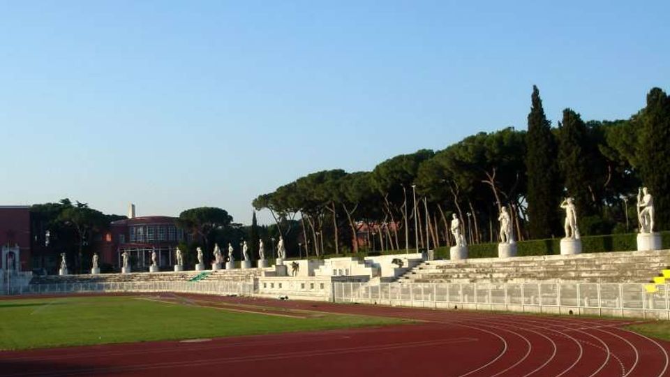 Stadio all’aperto con pista di atletica rossa che curva attorno al campo in erba e gradinate in pietra. Lungo le tribune sono allineate statue su piedistalli, con alberi alti e un edificio sullo sfondo sotto un cielo sereno.