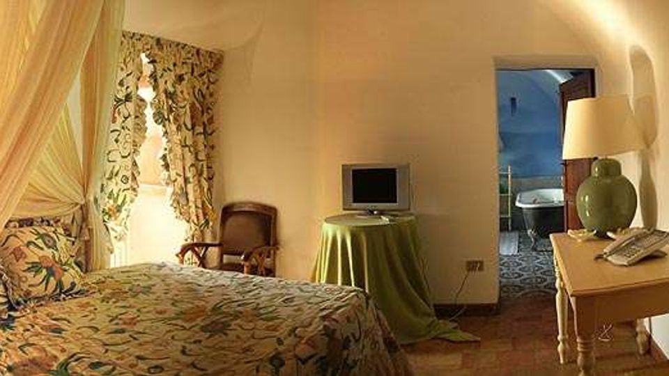 Camera da letto in stile rustico con letto a baldacchino e tessuti floreali, pareti bianche e pavimento in cotto. Un tavolino con lampada e telefono, una piccola TV su tavolo e un passaggio ad arco che conduce a un bagno con piastrelle decorative.