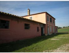 Casale di campagna con facciata rosa e tetto in tegole, composto da un corpo basso con finestre e da un edificio a due piani con persiane e camino. Intorno si vede un prato con sentiero ghiaioso e campi aperti sotto un cielo sereno.