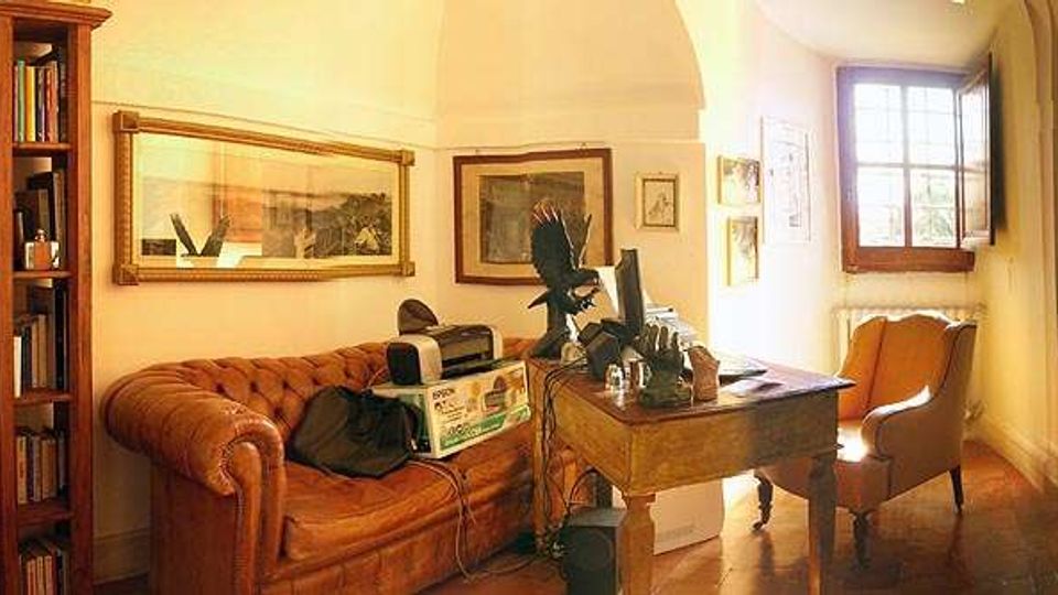 Interno di uno studio domestico con divano in pelle capitonné, libreria alta in legno, scrivania con computer e oggetti decorativi, e una poltrona vicino a una grande finestra. Pareti con quadri incorniciati e pavimento in legno in una stanza illuminata da luce naturale.