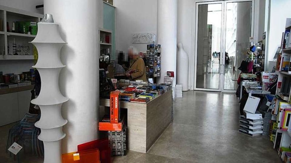 Interno di una libreria o negozio di libri moderno con pavimento in cemento, grandi colonne bianche e scaffali pieni di volumi e riviste. Al centro c’è un bancone basso con pile di libri, mentre sul fondo si vedono porte a vetri che conducono a un corridoio luminoso.