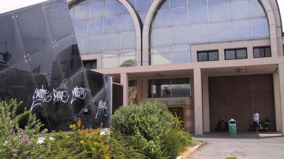 Facciata di un edificio moderno con grandi vetrate e due ampie arcate superiori; l’ingresso è sotto un portico con pilastri. In primo piano ci sono aiuole con arbusti e fiori e, sulla sinistra, una struttura angolare rivestita di pannelli scuri con graffiti.