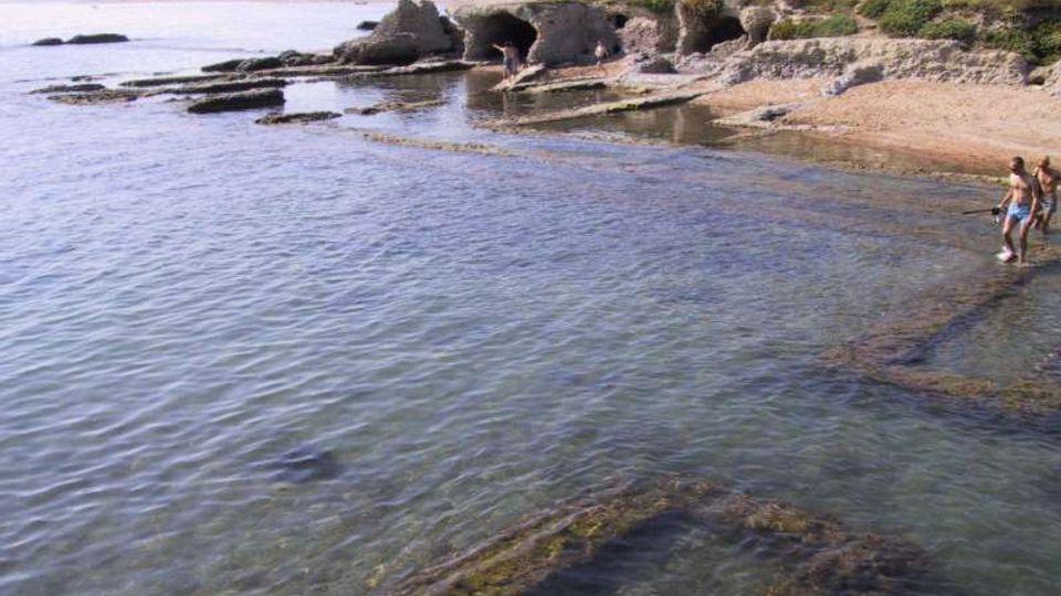 Costa rocciosa con piccole calette e acqua bassa e limpida, con scogli affioranti e piattaforme di roccia coperte di alghe. Sullo sfondo si vedono grotte nella falesia, vegetazione costiera e un edificio vicino alla scogliera.