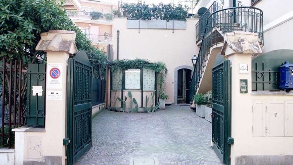 Ingresso con cancello in ferro battuto che conduce a un cortile interno pavimentato in sampietrini, con pareti chiare e vegetazione rampicante. Sulla destra si vede una scala esterna in metallo con balconcino, e sullo sfondo un edificio con portone ad arco e piccolo spazio verde.