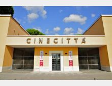 Ingresso degli Studi di Cinecittà, con facciata simmetrica color crema e ocra e grande scritta “CINECITTÀ” in alto. Al centro c’è un portone scuro affiancato da due banner verticali, con ampie cancellate metalliche ai lati e cielo azzurro con nuvole sullo sfondo.