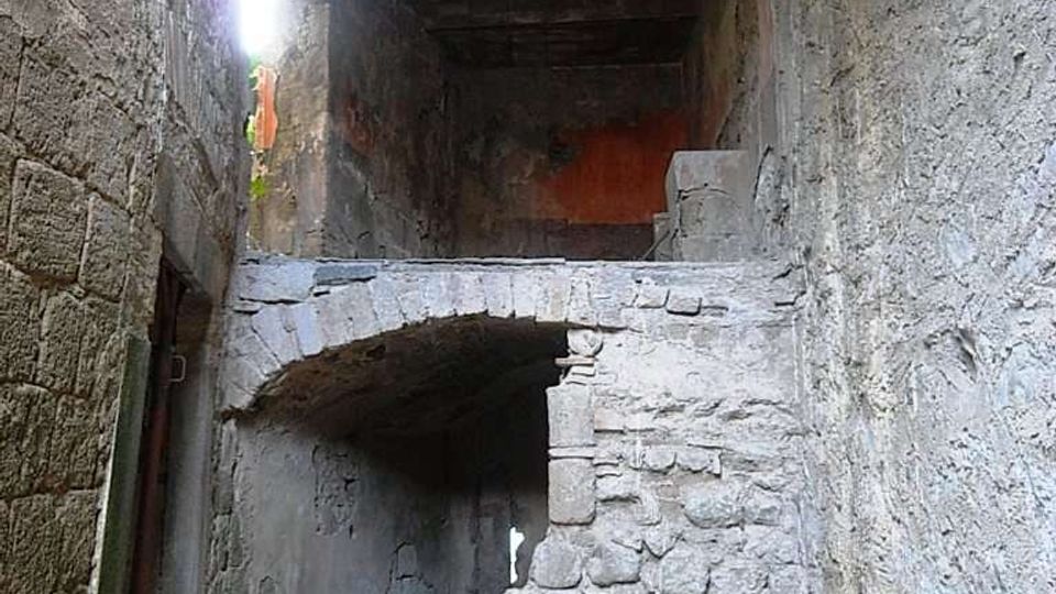 Interno di un edificio in pietra in rovina, con un arco in muratura parzialmente crollato e macerie sul pavimento. Pareti e soffitto grezzi e danneggiati, illuminati da una piccola apertura laterale.