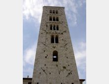 Torre medievale in pietra a pianta rettangolare, ripresa dal basso, con più livelli di finestre ad arco e aperture geminate sovrapposte. La facciata presenta mensole e tiranti metallici visibili, con cielo parzialmente nuvoloso sullo sfondo.