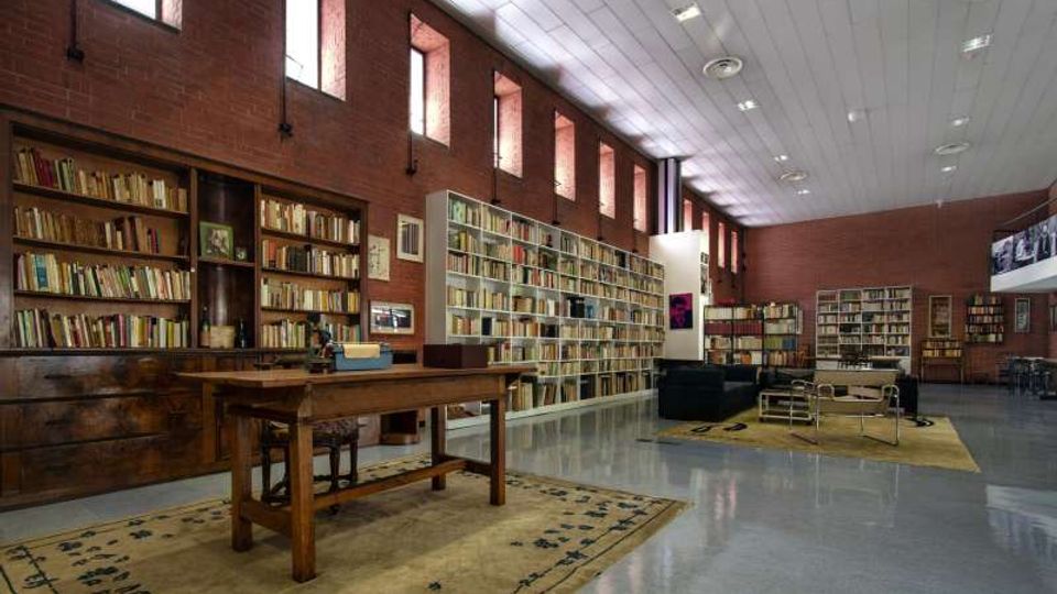 Interno di una biblioteca con pareti in mattoni rossi, alte librerie piene di libri e finestre strette in alto che lasciano entrare luce. In primo piano c’è un grande tavolo in legno su un tappeto, con area lettura sullo sfondo e soffitto alto con illuminazione a pannelli.