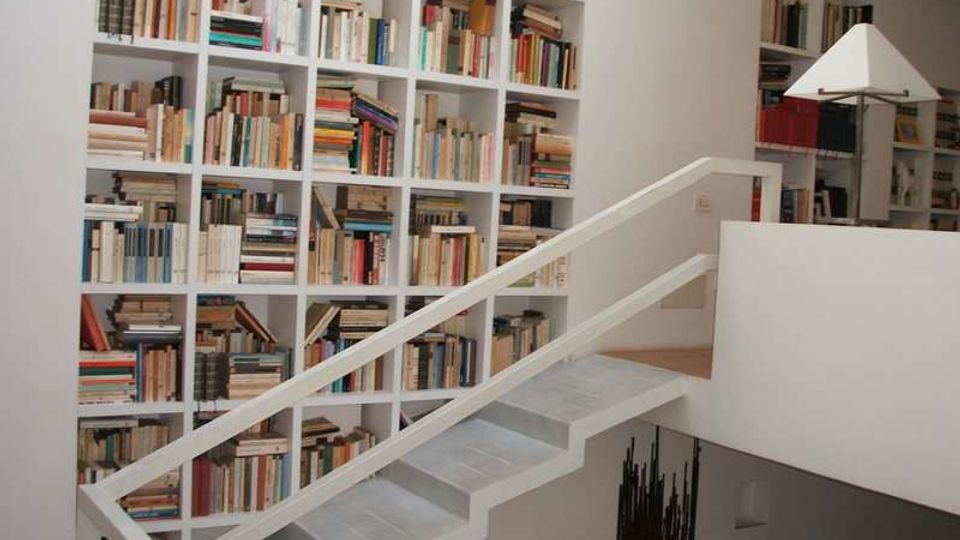 Interno di una casa con grande libreria a parete composta da scaffali bianchi a griglia pieni di libri. Una scala moderna bianca con ringhiera attraversa l’immagine e collega a un soppalco con parapetto.