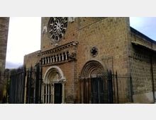 Facciata in pietra di una chiesa medievale con grande rosone circolare, portali ad arco decorati e piccoli colonnati scolpiti. Davanti all’ingresso sono presenti alte cancellate e recinzioni in ferro battuto.