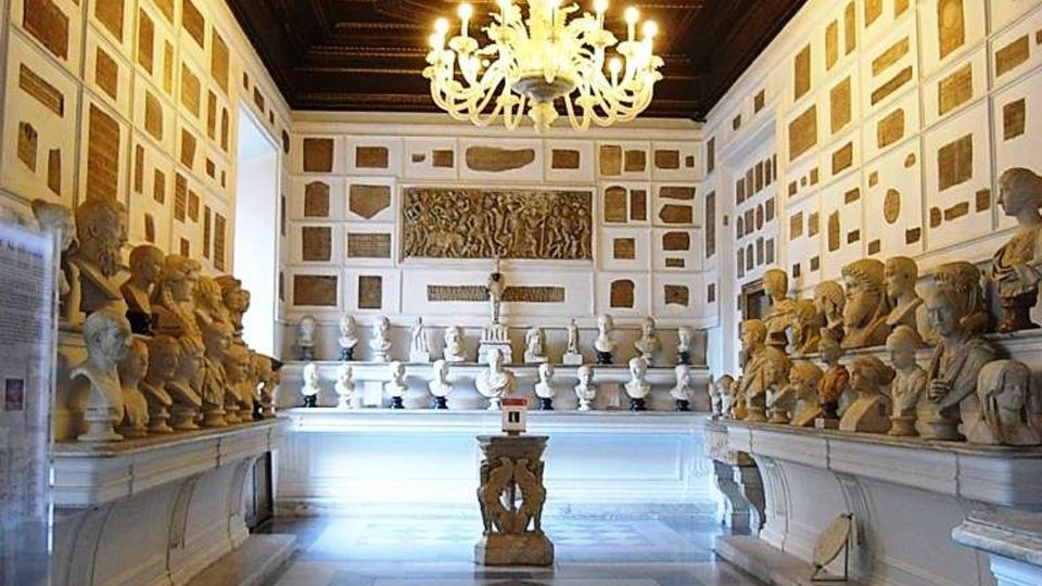 Interno di una galleria museale con soffitto in legno a cassettoni e grande lampadario centrale, pavimento in marmo a motivi geometrici. Lungo le pareti bianche con riquadri decorativi sono disposti numerosi busti e statue su mensole, con un piedistallo al centro della sala.