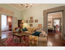 Soggiorno in stile classico con divani beige, tavolino basso e grande tappeto rosso su pavimento in parquet. Pareti verde chiaro con quadri incorniciati, lampadario a soffitto e ampie porte che collegano a stanze adiacenti con arredi coordinati.