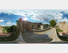 Panorama a 360 gradi di un complesso fortificato in rovina con mura e torri in pietra, affacciato su una valle alberata. Un camminamento in pietra con parapetti conduce tra edifici bassi e resti delle mura, con un grande albero e cielo parzialmente nuvoloso sullo sfondo.