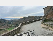 Terrazza panoramica lastricata in pietra con muretto di contenimento, affiancata da un edificio in muratura e una ringhiera metallica. Sullo sfondo si apre una vista su colline boscose, campi nella valle e un lago in lontananza sotto un cielo nuvoloso.
