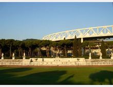 Campo sportivo con pista e prato verde in primo piano, delimitato da una bassa recinzione e da una terrazza con statue in pietra. Sullo sfondo si vedono alberi alti e una grande struttura bianca a traliccio simile a uno stadio sotto un cielo sereno.
