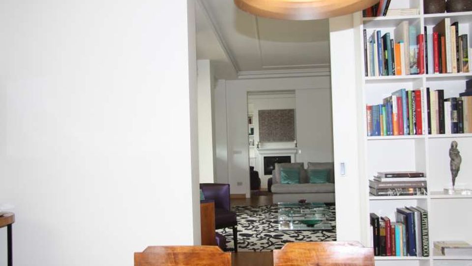 Interno di un appartamento moderno con pareti bianche, libreria a tutta altezza piena di libri e lampada a sospensione sopra un tavolo in legno. Sullo sfondo si vede un soggiorno con divano grigio, tavolino in vetro, tappeto a motivi in bianco e nero e camino.