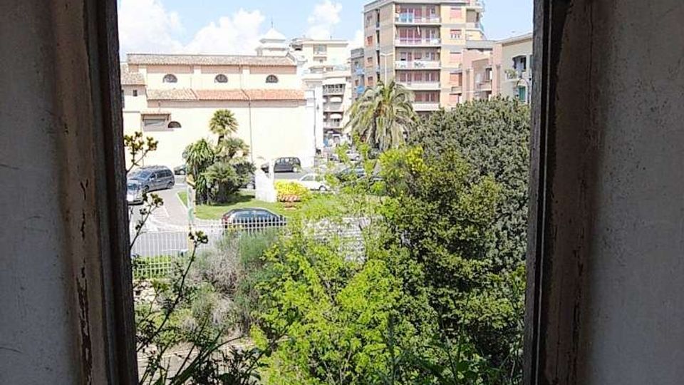 Vista attraverso una piccola finestra quadrata con cornice in cemento, da un interno spoglio, su un’area verde con alberi e palme. Sullo sfondo si vedono parcheggio e palazzine residenziali a più piani sotto un cielo parzialmente nuvoloso.