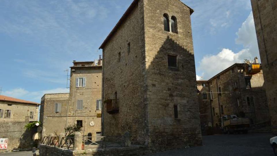 Torre in pietra in un borgo storico, con piccole finestre e apertura a bifora nella parte superiore. Intorno si vedono edifici in muratura e una piazzetta con pavimentazione in pietra sotto un cielo sereno.