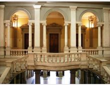 Interno di un edificio storico con loggiato a due livelli, archi a tutto sesto e colonne in pietra. Al centro una balconata con balaustra e scale laterali conduce a un corridoio con porte in legno illuminato da luci calde.