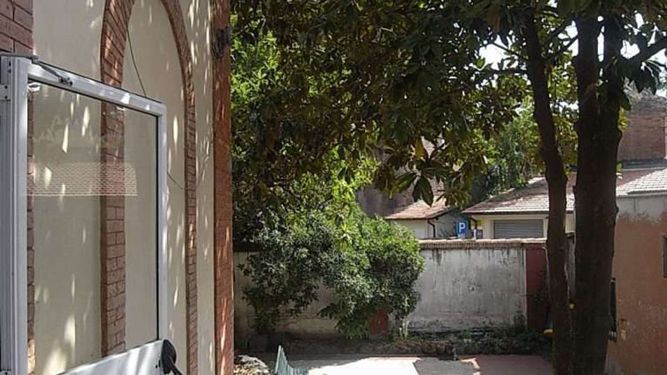 Cortile esterno pavimentato accanto a un edificio in muratura con arco in mattoni, con scala che scende e corrimano metallico. Alberi alti ombreggiano l’area; sullo sfondo si vedono un muretto, vegetazione e alcuni edifici bassi.