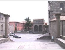 Piazza lastricata in un borgo storico, vista dall’alto di una scalinata in pietra con due colonne in primo piano. Attorno si vedono edifici in muratura con tetti in coppi, un porticato con archi, un grande albero centrale e alcune auto parcheggiate.